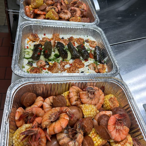 Three foil pans filled with shrimp, corn, potatoes, sausage, and stuffed grape leaves with sauce.