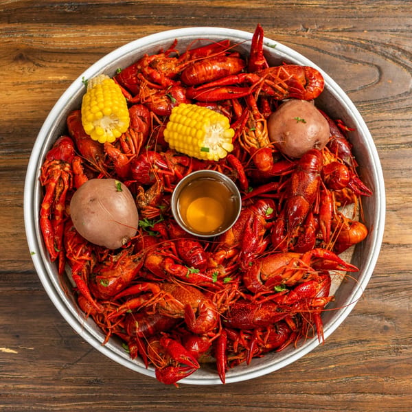 A large metal platter overflowing with bright red crawfish, corn on the cob, and potatoes, with a small cup of dipping sauce.