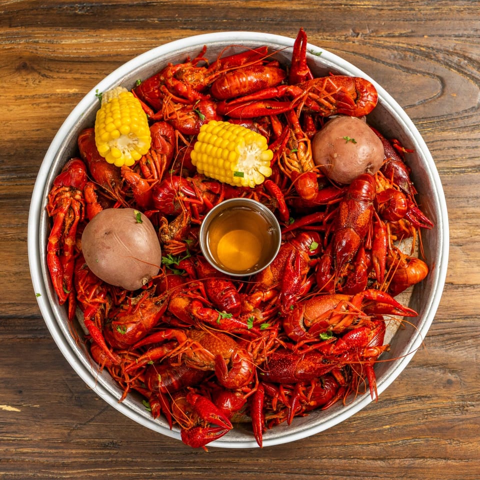 A large metal platter overflowing with bright red crawfish, corn on the cob, and potatoes, with a small cup of dipping sauce.