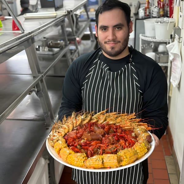 Chef holding a large platter of crawfish, shrimp, corn on the cob, and sausage in a kitchen.