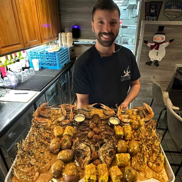 A smiling man holds a large tray of Cajun seafood boil with crab, shrimp, fish, corn, and potatoes.
