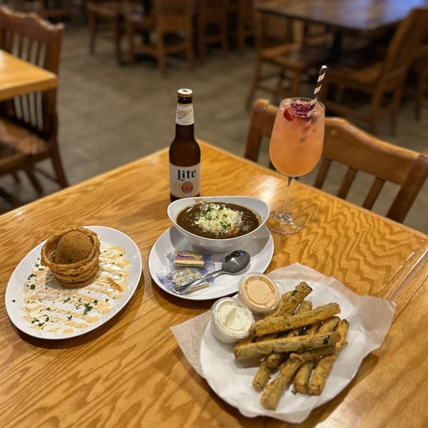 A table with a bowl of gumbo, fried okra, a blooming onion, a beer, and a cocktail.