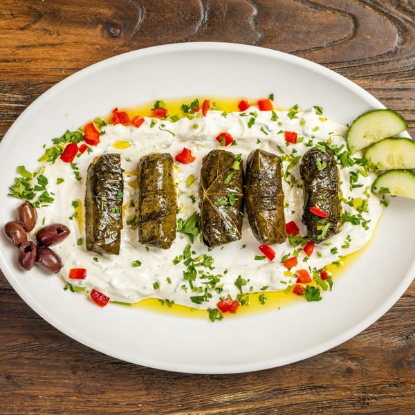 Five dolmades on a white plate with yogurt sauce, red peppers, parsley, olives, and cucumber.