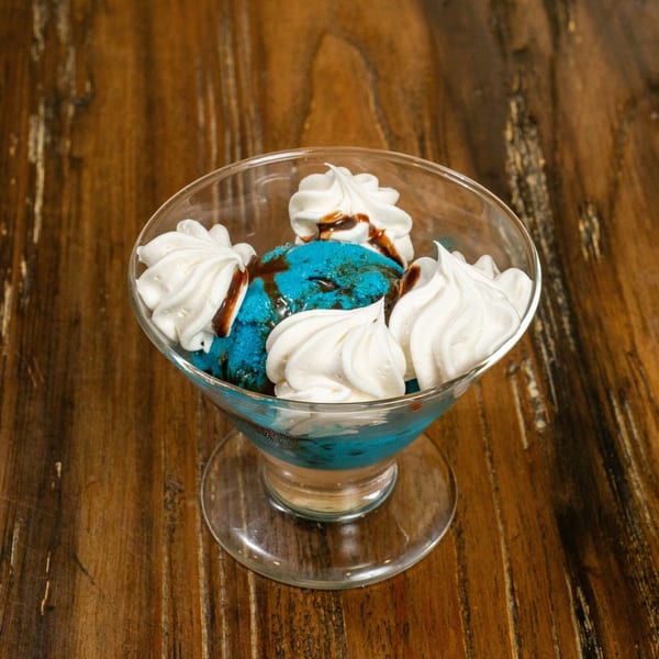 Blue ice cream with chocolate syrup and whipped cream in a glass dish on a wooden table.