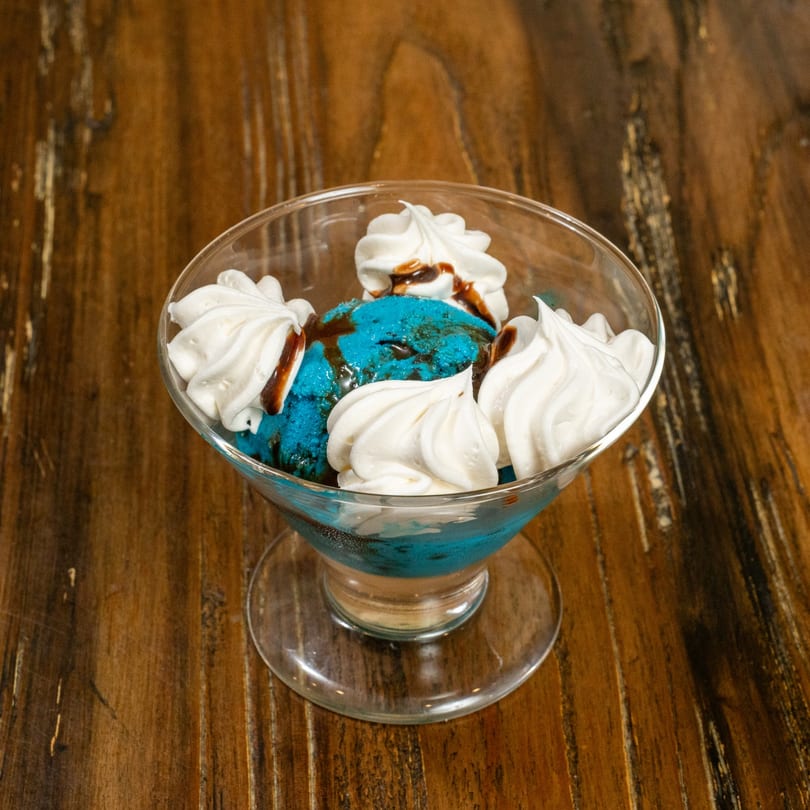 Blue ice cream with chocolate syrup and whipped cream in a glass dish on a wooden table.