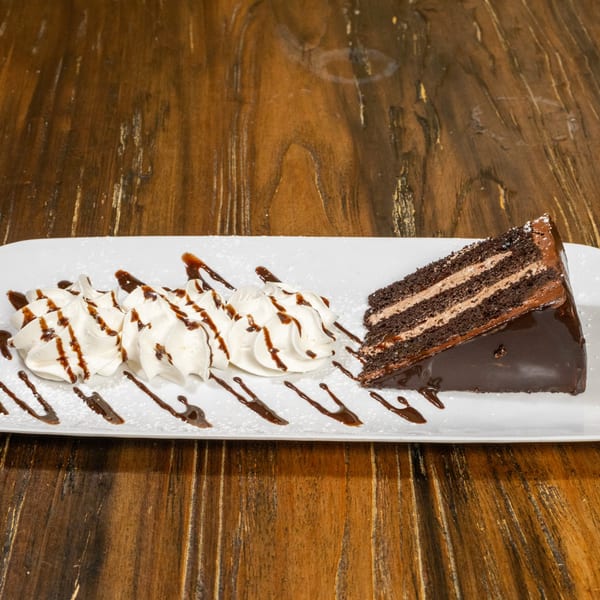 Slice of chocolate cake with whipped cream and chocolate drizzle on a white plate.