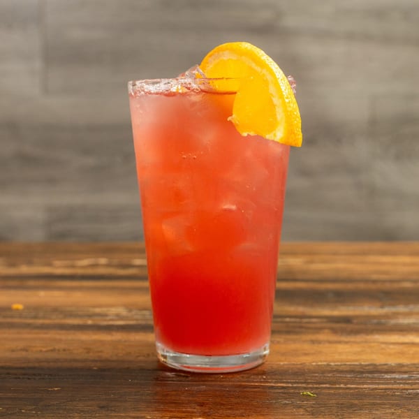 Red cocktail with ice and an orange slice garnish, on a wooden table.