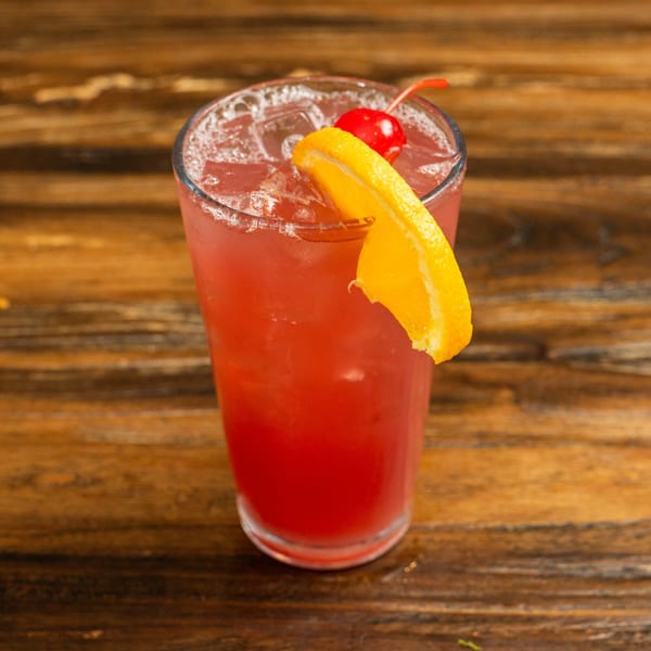 Red cocktail with ice, garnished with an orange slice and a maraschino cherry on a wooden table.