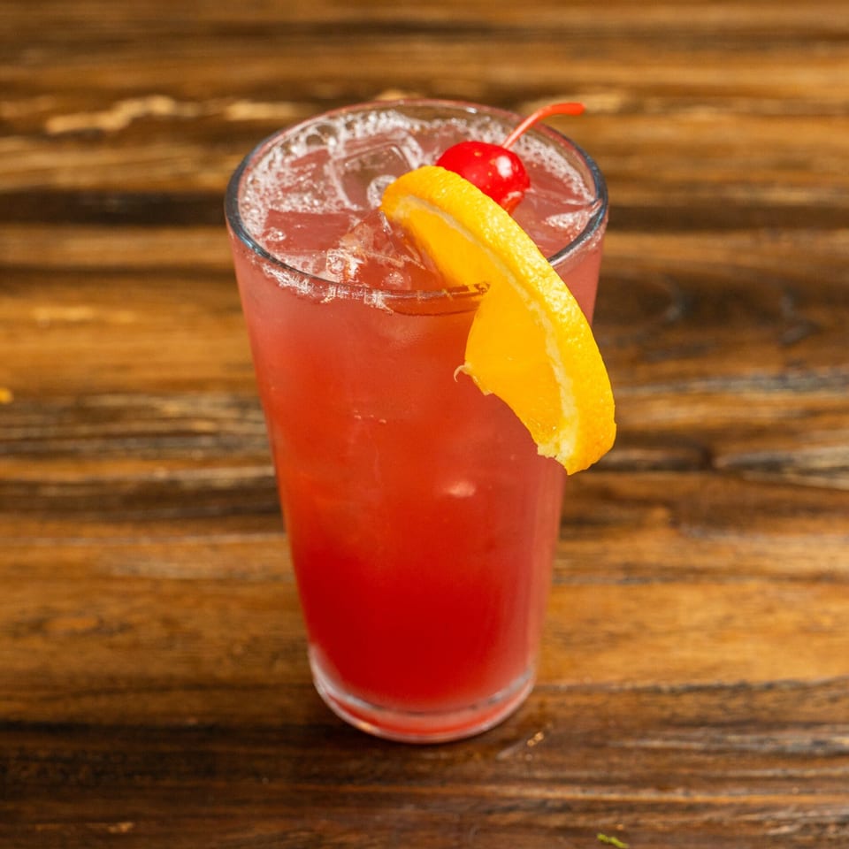 Red cocktail with ice, garnished with an orange slice and a maraschino cherry on a wooden table.