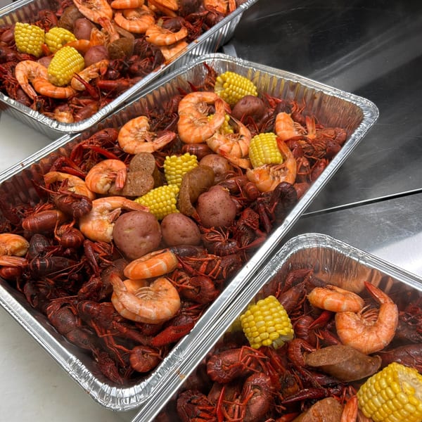 Three foil pans filled with a seafood boil of crawfish, shrimp, corn on the cob, potatoes, and sausage.