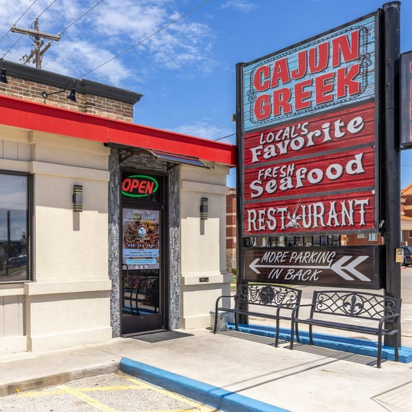 Exterior of Cajun Greek restaurant with a large sign, benches, and an open door.