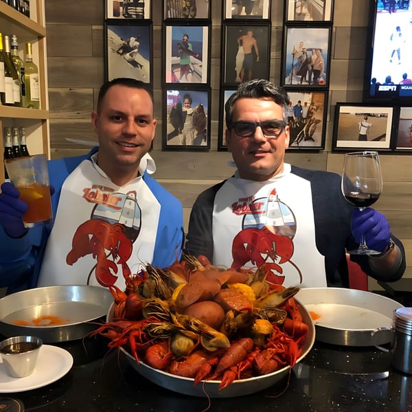 Two men in lobster bibs and gloves smile, holding drinks over a large bowl of seafood boil.