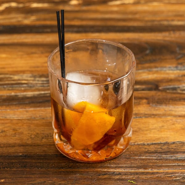 A cocktail with ice, an orange peel, and two black straws on a wooden table.