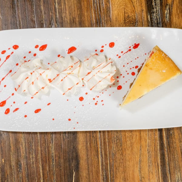 A slice of cheesecake with whipped cream and strawberry drizzle on a white rectangular plate.