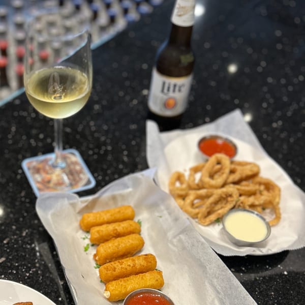 Mozzarella sticks, onion rings, white wine, and a Miller Lite beer on a dark bar top.