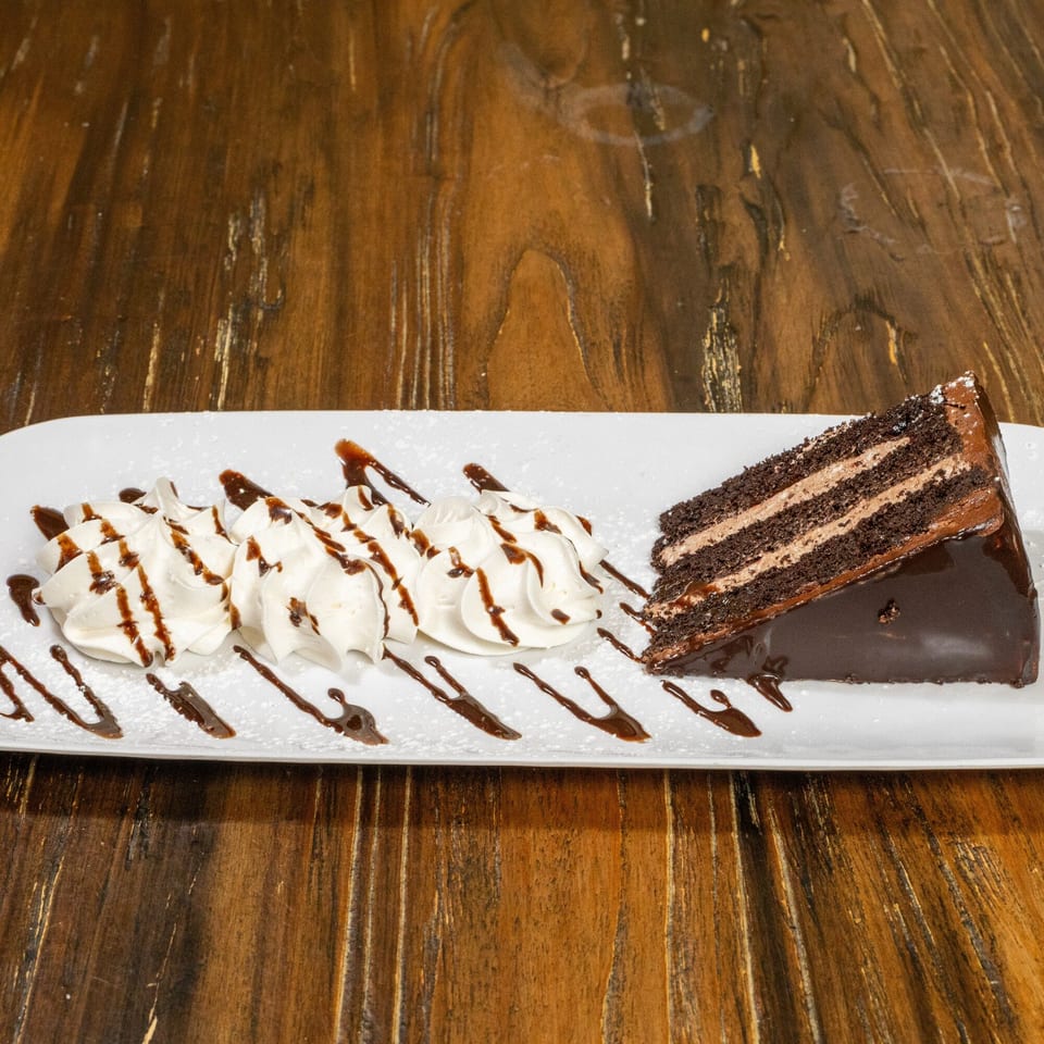 A slice of chocolate cake with whipped cream and chocolate drizzle on a white plate.