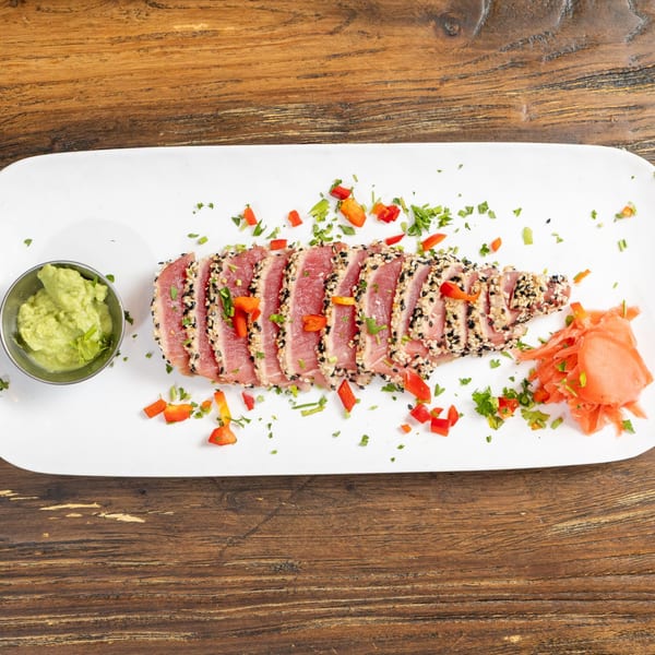 Seared tuna with sesame seeds, chopped peppers, cilantro, wasabi, and pickled ginger on a white plate.