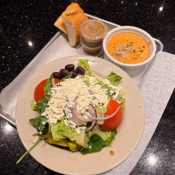 A Greek salad with feta, olives, and tomatoes, alongside soup, bread, and dressing on a tray.