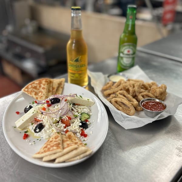 Greek salad with pita, fried calamari with sauce, and two beer bottles on a metal table.
