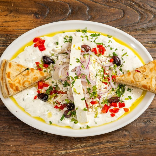 A white platter of creamy dip topped with feta, olives, red peppers, onions, and pita bread.