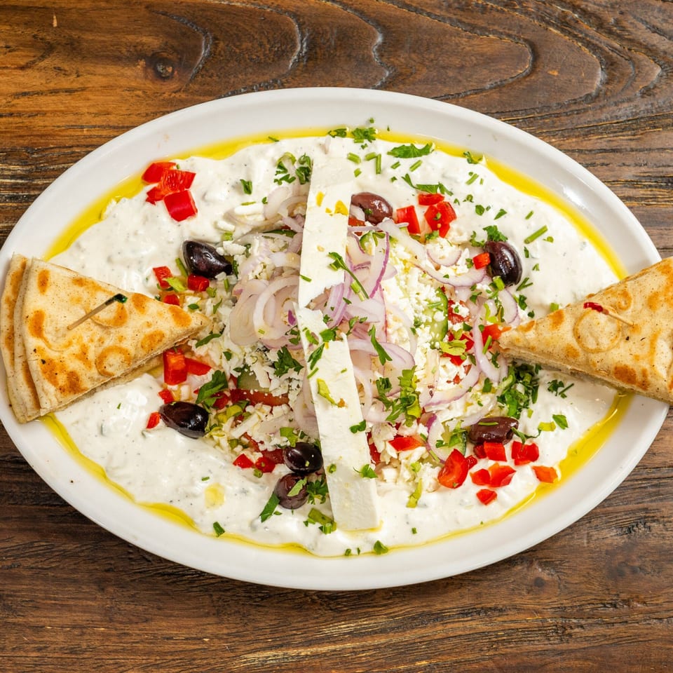 A white platter of creamy dip topped with feta, olives, red peppers, onions, and pita bread.