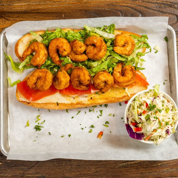 A shrimp po'boy sandwich with lettuce and tomatoes, served with a side of coleslaw.