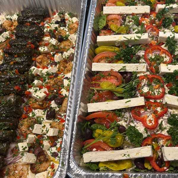 Two foil trays of Greek food, one with dolmades and shrimp, the other with salad and feta.