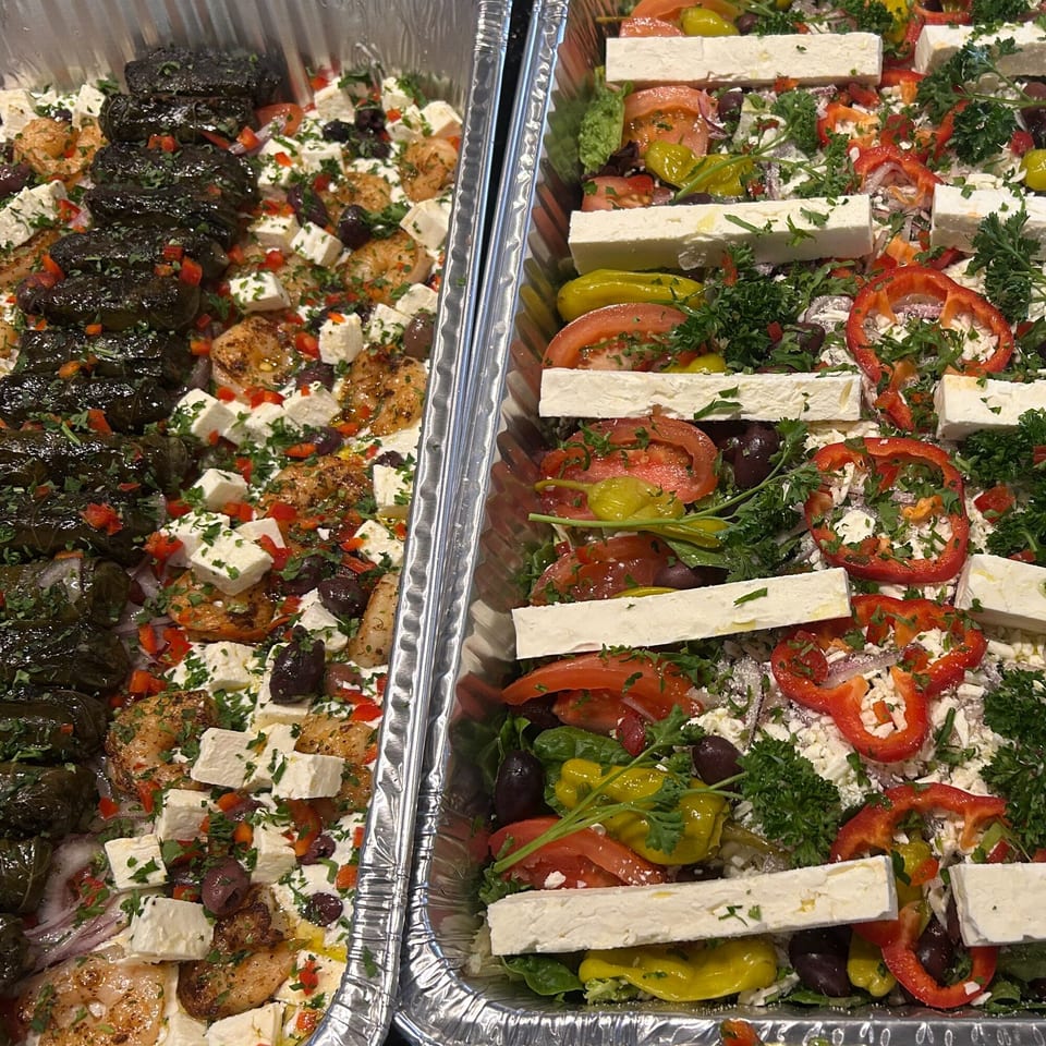 Two foil trays of Greek food, one with dolmades and shrimp, the other with salad and feta.