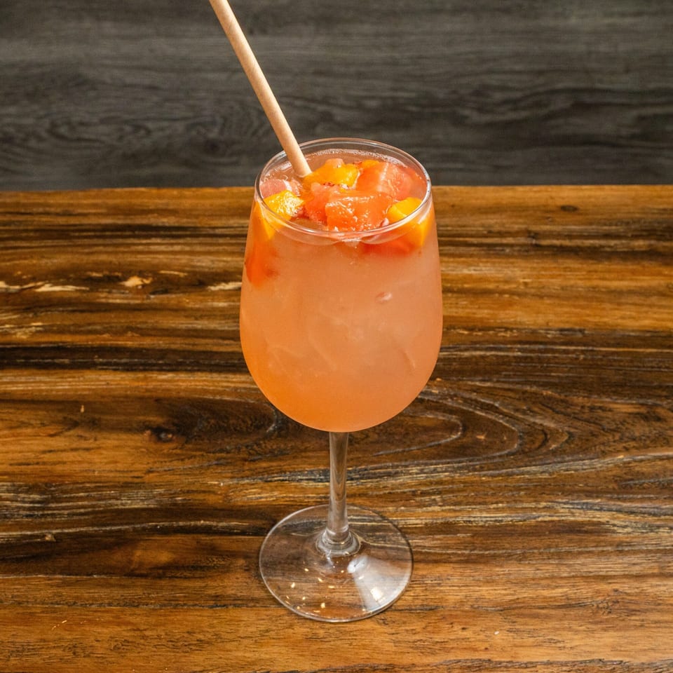 A refreshing pink cocktail with fruit chunks and a straw, on a rustic wooden table.