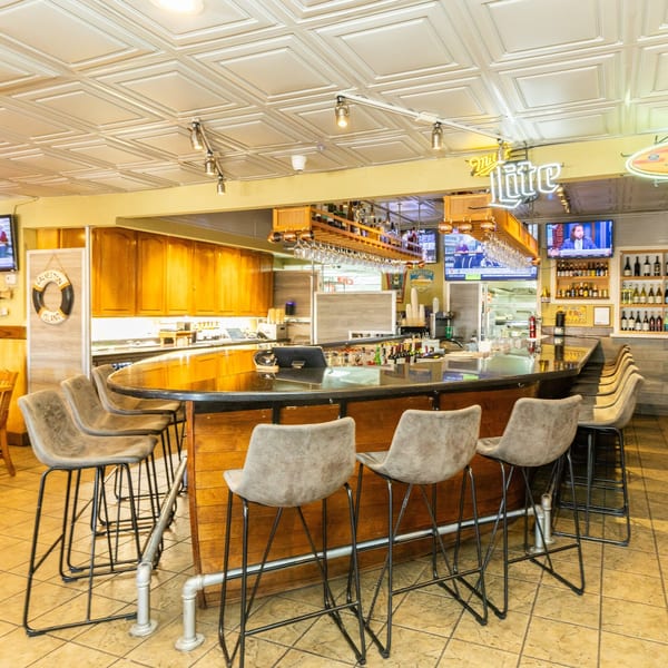 A well-lit, empty restaurant bar with a dark countertop, bar stools, and a Miller Lite neon sign.