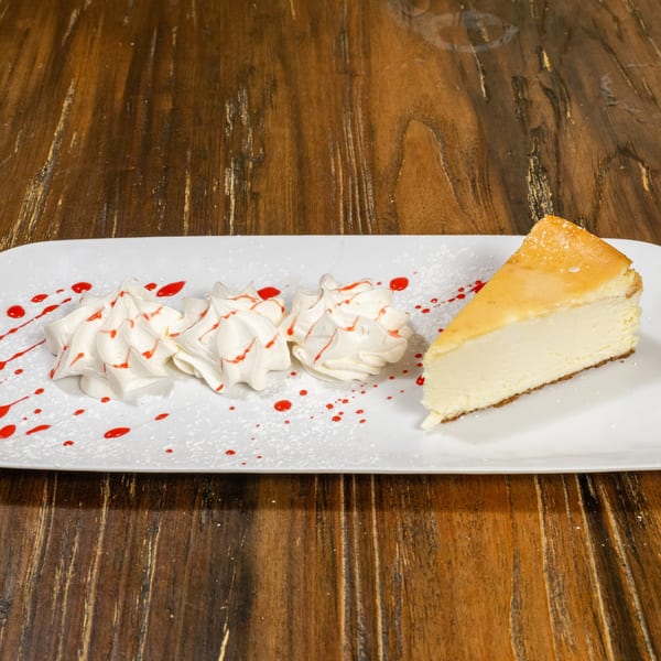 A slice of cheesecake with whipped cream and raspberry drizzle on a white rectangular plate.