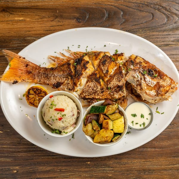 Grilled whole fish with charred lemon, mashed potatoes, roasted vegetables, and a side of sauce.