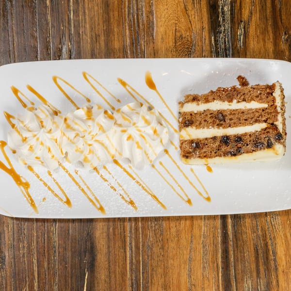 A slice of carrot cake with cream cheese frosting, whipped cream, and caramel drizzle on a white plate.