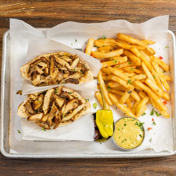 Two chicken shawarma sandwiches, a pile of french fries, a pepper, and a dipping sauce on a tray.