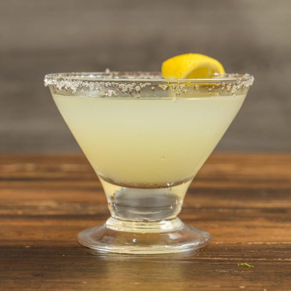 A lemon margarita in a salt-rimmed glass with a lemon wedge on a wooden table.