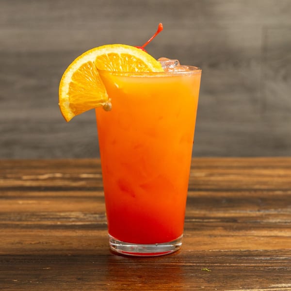 A vibrant orange and red cocktail, garnished with an orange slice and cherry, on a wooden table.