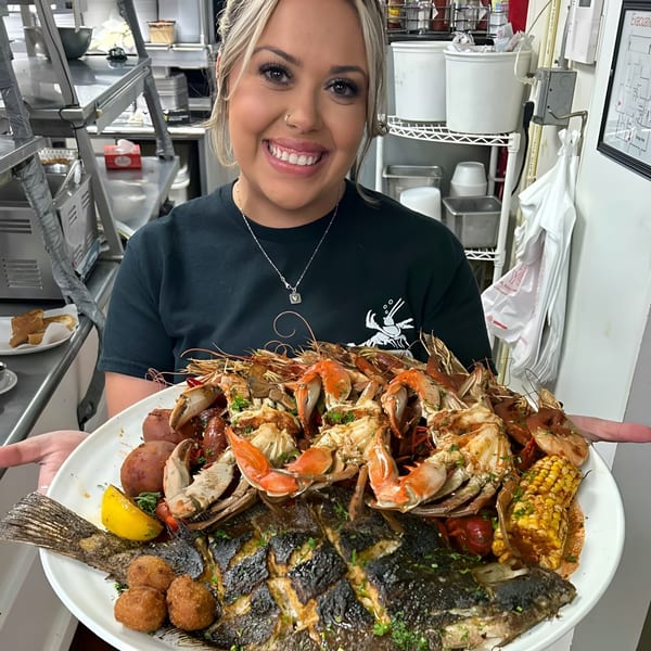 Smiling server holding a large platter of grilled fish, crab, shrimp, corn, and potatoes in a kitchen.