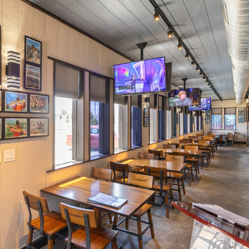 A long, narrow dining room with wooden tables and chairs, TVs, and framed pictures on the walls.