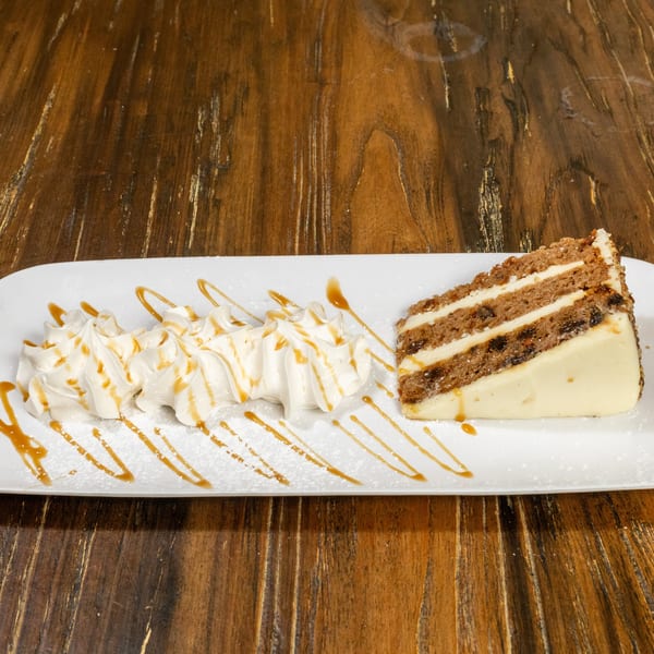 A slice of layered cake with cream cheese frosting and whipped cream drizzled with caramel.