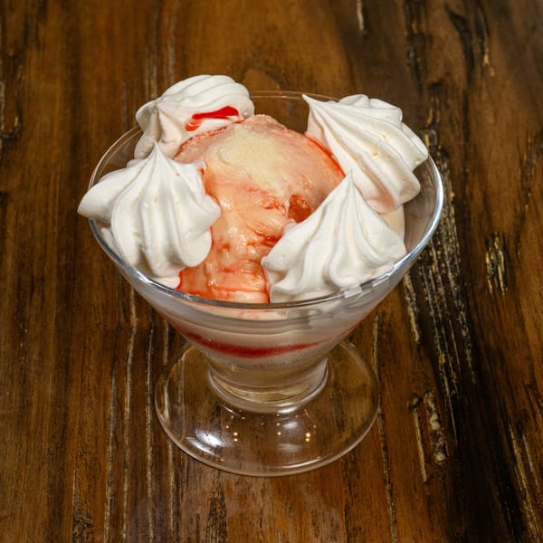 Pink ice cream with white meringue swirls and red syrup in a glass bowl on a wooden table.