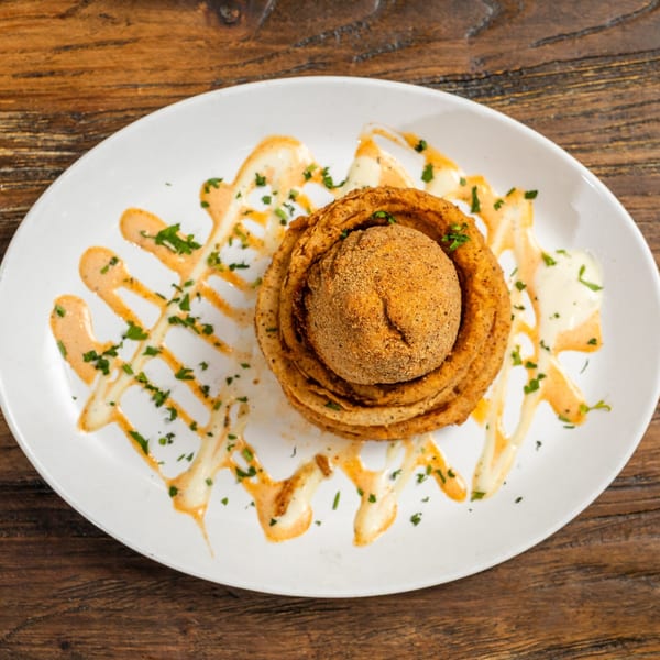 Crispy fried onion rings stacked around a breaded, spherical appetizer, drizzled with sauces and herbs.