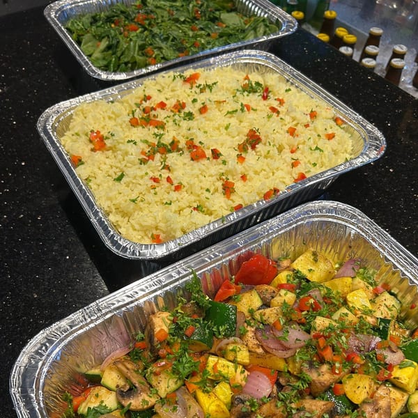 Three foil trays of food: green salad, yellow rice, and roasted vegetables, on a black countertop.