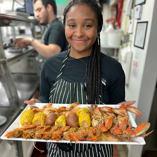 Smiling chef holding a large platter of seafood boil with shrimp, crab, corn, and potatoes.