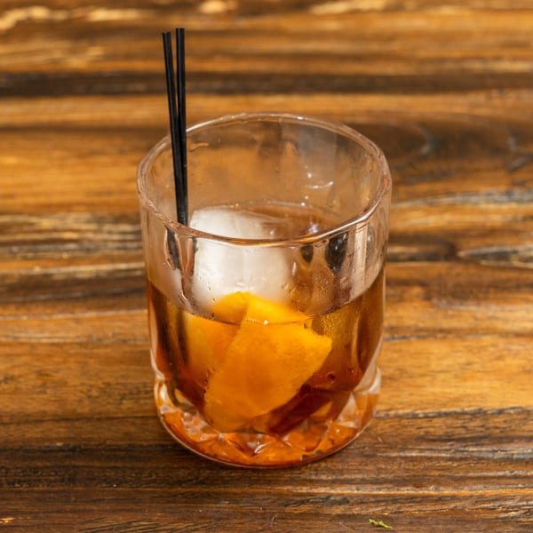 An Old Fashioned cocktail with a large ice cube, orange peel, and two black straws on a wooden table.